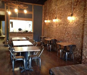 Restaurant interior with metal chairs and tables against a brick wall, illuminated by pendant lights.