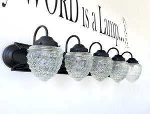 Four-light vanity fixture with clear glass shades, black metal frame, mounted on a white wall.