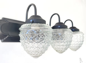 Three-light vanity fixture with clear glass shades featuring a textured acorn design and matte black metal accents.