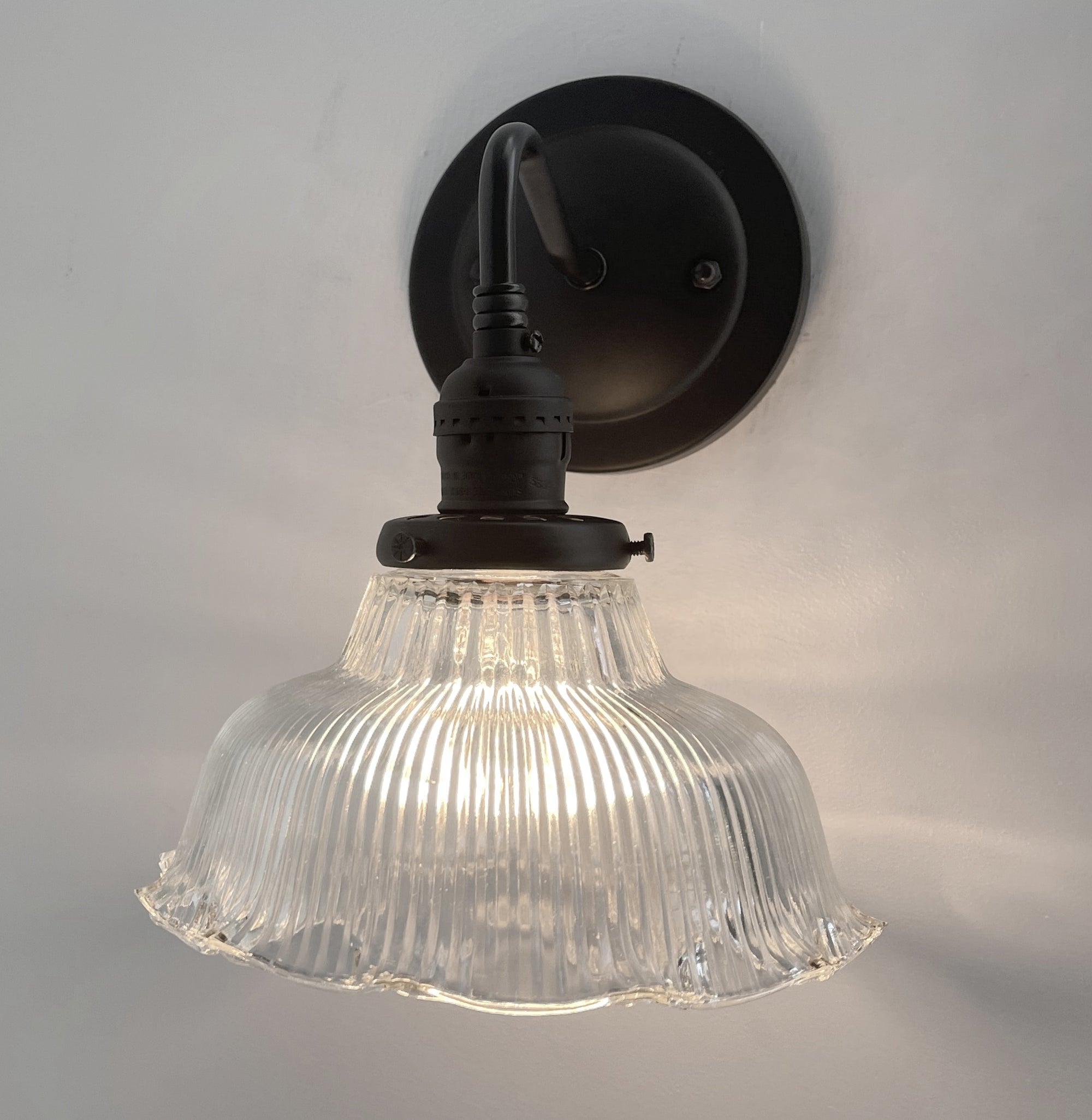 Clear glass ribbed wall sconce with a black metal mount, illuminating a light grey wall.
