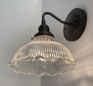Clear glass wall sconce with ribbed detailing and scalloped edge, mounted on a dark metal arm against a light wall.
