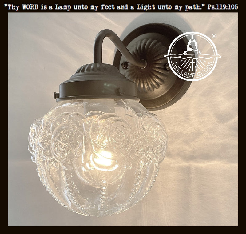 Clear glass globe lamp shade with embossed rose pattern, mounted on a dark metal wall sconce.
